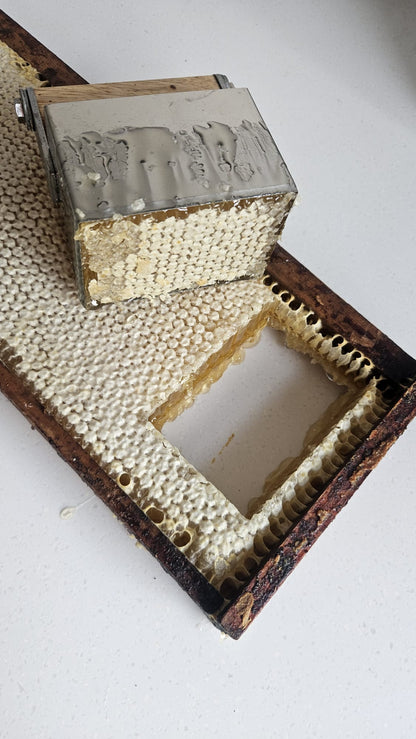 A close-up of a wooden beekeeping frame with honeycomb cells, a metal lid, and a rectangular section removed or opened, photographed on a plain light background.
