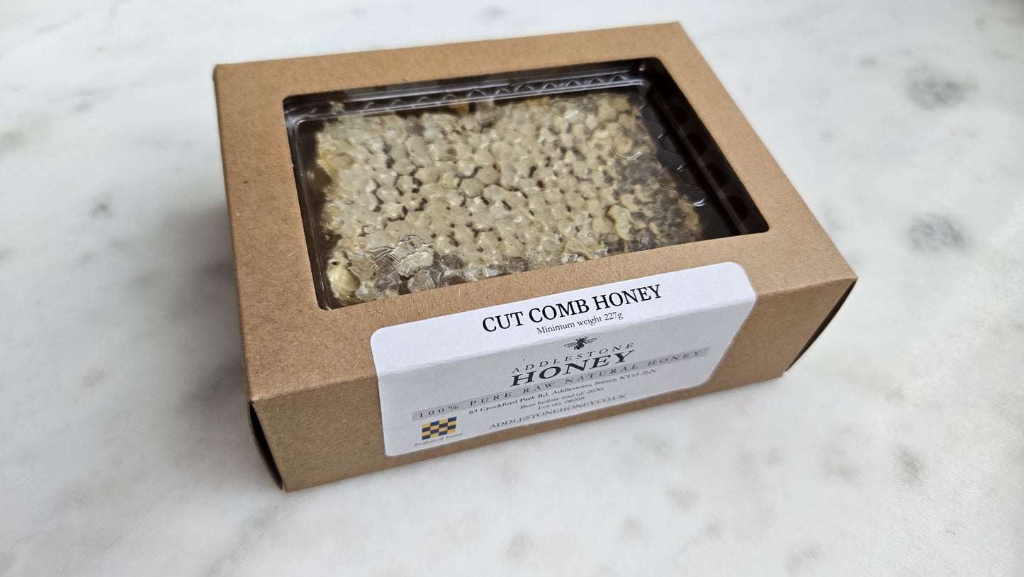 A square brown kraft cardboard box of honey with a clear window showing the honeycomb structure inside, and a white label reading 'CUT COMB HONEY' on a light marble surface.