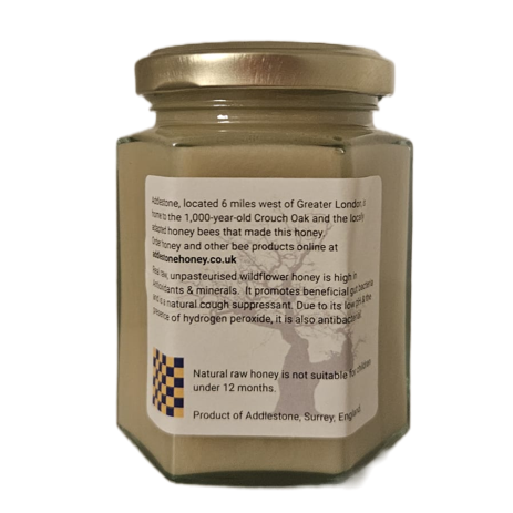 A hexagonal glass jar of Addlestone raw wildflower creamed honey, with a metal lid and a white label showing product details, isolated on a white background.
