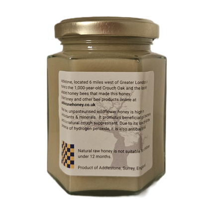 A hexagonal glass jar of Addlestone raw wildflower creamed honey, with a metal lid and a white label showing product details, isolated on a white background.